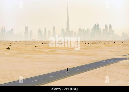Splendida vista aerea di una persona non identificata che cammina su una strada deserta coperta da dune di sabbia con lo skyline di Dubai sullo sfondo. Foto Stock