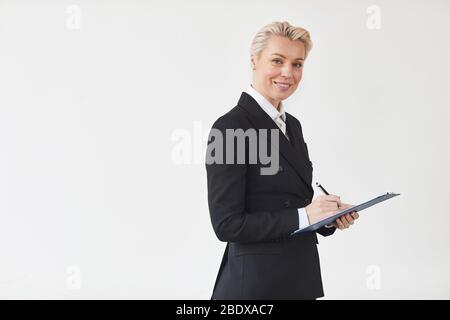 Ritratto di donna d'affari matura in abito nero prendere appunti in documento e sorridere alla macchina fotografica su sfondo bianco Foto Stock