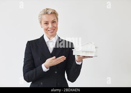 Maturo realtor in abito nero che tiene modello della casa e sorridente a macchina fotografica isolato su sfondo bianco Foto Stock