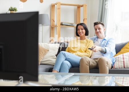 Giovane coppia sorridente seduta sul divano e guardando la TV nel soggiorno a casa Foto Stock