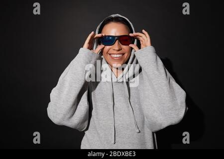Ritratto di giovane donna felice con cappuccio indossando occhiali 3D e guardando film isolato su sfondo nero Foto Stock