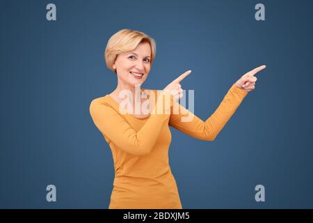 Stile libero. Donna matura in piedi isolato su blu puntando da parte al copy space sorridente allegro Foto Stock
