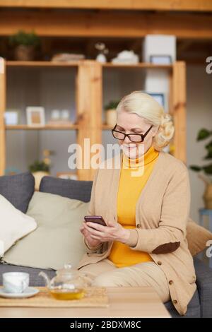 Donna anziana seria negli occhiali che usano il suo telefono cellulare mentre riposava sul divano a casa Foto Stock