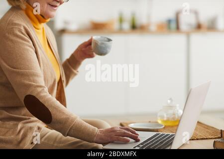 Primo piano di una donna matura che tiene una tazza di tè e digita sul computer portatile mentre si siede sul divano a casa Foto Stock