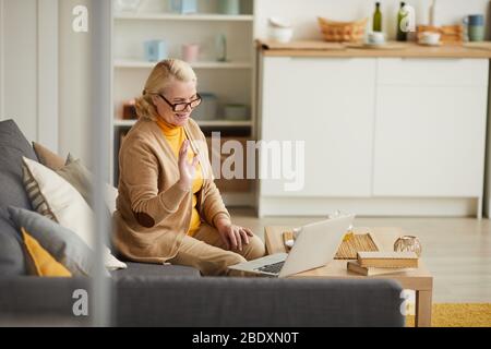 Felice donna matura in occhiali che agita la mano e comunica online sul laptop mentre si siede sul divano nel soggiorno Foto Stock