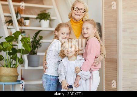 Ritratto di nonna felice in occhiali abbracciando i suoi nipoti e sorridendo alla macchina fotografica mentre stanno in piedi nella stanza a casa Foto Stock