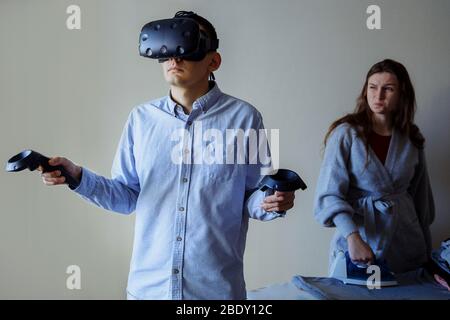Un uomo con una camicia blu in denim gioca in realtà virtuale. Moglie che strofinava le camicie e gli sguardi offesi al marito Foto Stock