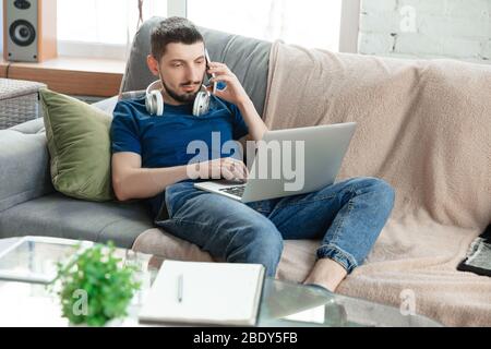 Giovane uomo concentrato studiare a casa durante corsi online o informazioni gratuite da hisself. Diventa sviluppatore di software, responsabile IT mentre è isolato, in quarantena. Utilizzo di laptop, smartphone, cuffie. Foto Stock