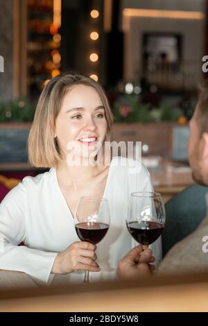 Bella giovane donna con un sorriso sooty che parla con il suo ragazzo mentre fa brindisi con bicchieri di vino rosso in ristorante di classe Foto Stock