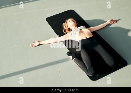 Ragazza piuttosto attiva in tuta sdraiata sul tappetino nero con le sue braccia distese e le gambe piegate in ginocchia, mentre si pratica l'esercizio di relax Foto Stock