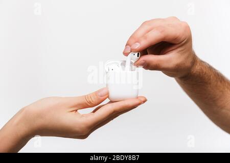 Immagine di primo piano delle mani di un uomo e di una donna isolata su sfondo bianco da parete che contiene auricolari wireless bluetooth. Foto Stock