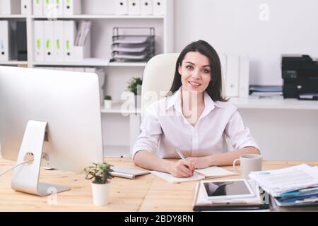 Felice giovane bruna donna d'affari in formalwear guardarsi mentre scrivono giù il programma di lavoro di giorno al mattino in ufficio Foto Stock