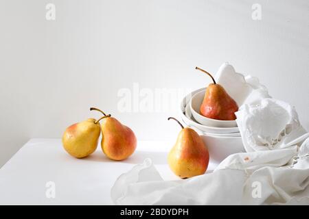 Ancora vita con duchessa di pera giallo-rossa in un mucchio di ciotole e un tovagliolo ricamato bianco sulla parete bianca. Spazio di copia Foto Stock