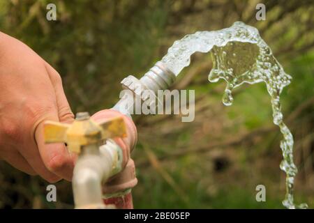 Acqua che scorre da un rubinetto di metallo esterno. Una mano sta chiudendo il rubinetto. Foto Stock