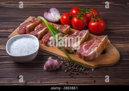 Fette crude di pancetta di maiale su un tagliere, su un tavolo rurale in legno. Pancia di maiale con verdure. Foto Stock
