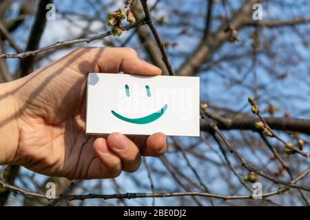 Felice sorriso verde emoji su carta bianca tenendo in mano su sfondo blu primavera sole cielo. Positivo umore tempo buono Foto Stock