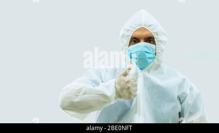 Medico in tuta bioprotettiva bianca e maschera blu in piedi con il braccio teso e la mano con il pollice in su facendo COME segno su sfondo bianco Foto Stock