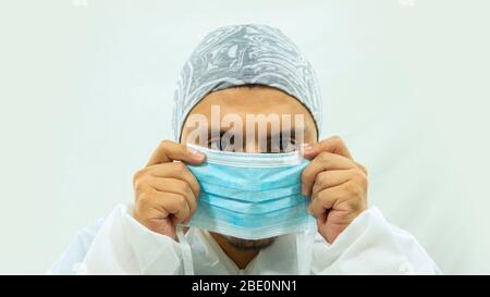 Medico in tuta bioprotettiva bianca con una maschera blu con le mani che la mette sul viso su sfondo bianco Foto Stock