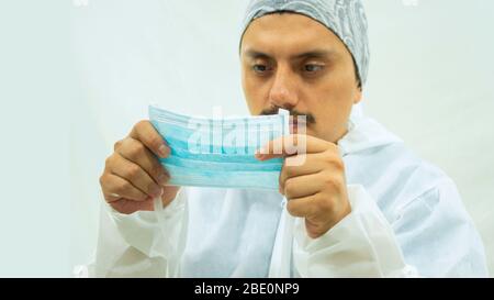 Medico in tuta bioprotettiva bianca che tiene una maschera blu con le mani in primo piano prima di metterla su sfondo bianco Foto Stock