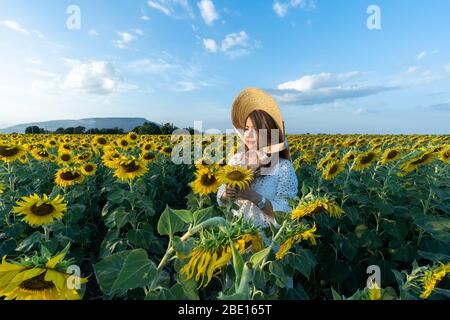 Una bella donna asiatica in un abito bianco e cappello a piedi su un campo di girasoli, sorridendo un bel sorriso, ragazza allegra, stile, stile di vita e sole Foto Stock