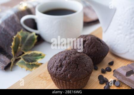 Muffin al cioccolato, patatine di cioccolato, una tazza di caffè, una tea pot su una superficie di legno con sciarpa calda, luci di Natale bokeh e agrifoglio nel Foto Stock