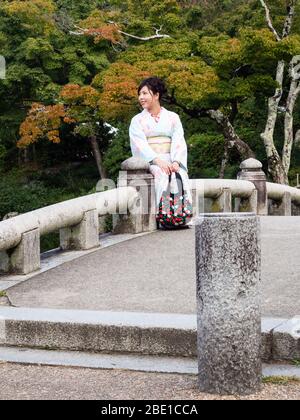 Kyoto, Giappone - 30 settembre 2015: Giovane ragazza in kimono bello seduto su un ponte di pietra in giardino giapponese tradizionale Foto Stock