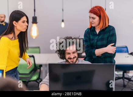 Due giovani donne d'affari accanto a un collega maschile mentre spiega le nuove attività online su un computer in un ambiente di ufficio confortevole. Foto Stock