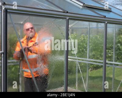 Addetto alle costruzioni che pulisce la sporcizia con pulitore ad alta pressione da una serra di vetro. Foto Stock