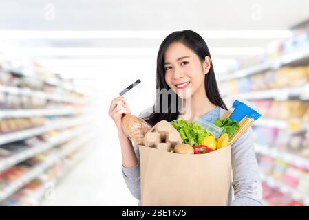 Bella donna asiatica che tiene sacchetto di carta pieno di acquisti alimentari con carta di credito in supermercato Foto Stock