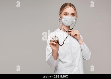 Giovane donna in uniforme medica e maschera protettiva utilizza uno stetoscopio e posa su sfondo grigio, isolato. Sistema sanitario e emergenza Foto Stock