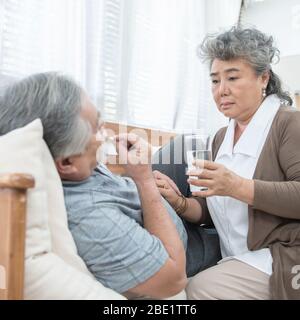 Uomo asiatico senior che prende medicinali e acqua potabile mentre si sdraia sul divano. La vecchia donna si prende cura del marito mentre la sua malattia a casa. Foto Stock