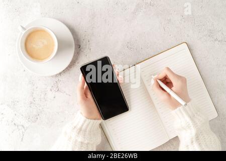 Belle mani da donna con smartphone aggiornato e scrittura in blocco note con tazza di caffè su sfondo concreto. Concetto di business femminile. COFF Foto Stock