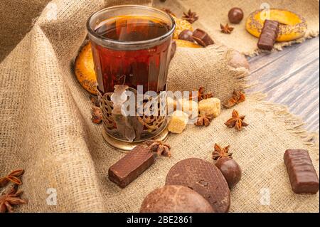 Un bicchiere sfaccettato di tè in un portabicchieri vintage, bagel, biscotti, cioccolato e torte al cioccolato su uno sfondo di tessuto grezzo casalinga. Primo piano Foto Stock