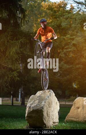 Ritratto di giovane ciclista in piedi sulla ruota posteriore di bici di bmx su una roccia guardando la macchina fotografica nel parco, alberi verdi sullo sfondo Foto Stock