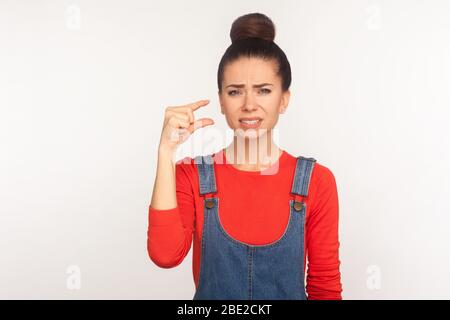 Troppo piccolo! Ritratto di ragazza insoddisfatta con capelli bun in tuta denim mostrando un po 'di gesto e guardando con espressione scettica delusa Foto Stock