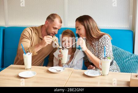 La famiglia felice sta facendo una pausa di milkshake nel caffè della città, mamma e papà stanno cercando di assaggiare il cocktail della figlia, trascorrendo del tempo insieme Foto Stock