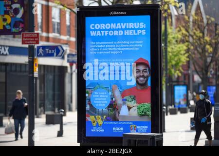 Watford, Regno Unito. 11 aprile 2020. Un uomo che indossa una maschera cammina da uno schermo pubblicitario nel centro commerciale di Watford il sabato di Pasqua durante il blocco come il coronavirus (COVID19) pandemia continua. La schermata mostra un messaggio del consiglio locale che promuove l'aiuto della comunità per coloro che ne hanno bisogno. Credit: Stephen Chung / Alamy Live News Foto Stock