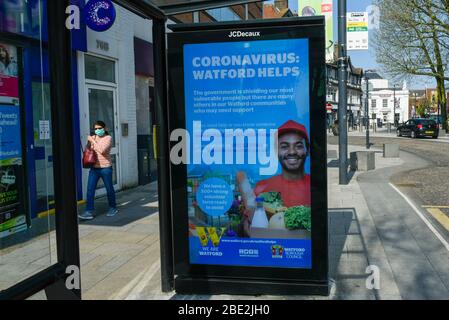 Watford, Regno Unito. 11 aprile 2020. Una donna che indossa un facemask passeggiate da uno schermo pubblicitario su un rifugio fermata autobus nel centro commerciale di Watford il Sabato di Pasqua durante il blocco come il coronavirus (COVID19) pandemia continua. La schermata mostra un messaggio del consiglio locale che promuove l'aiuto della comunità per coloro che ne hanno bisogno. Credit: Stephen Chung / Alamy Live News Foto Stock