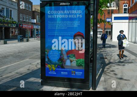 Watford, Regno Unito. 11 aprile 2020. Una donna che indossa un facemask passeggiate da uno schermo pubblicitario su un rifugio fermata autobus nel centro commerciale di Watford il Sabato di Pasqua durante il blocco come il coronavirus (COVID19) pandemia continua. La schermata mostra un messaggio del consiglio locale che promuove l'aiuto della comunità per coloro che ne hanno bisogno. Credit: Stephen Chung / Alamy Live News Foto Stock