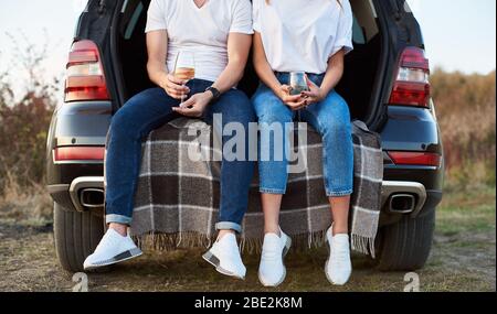 Primo piano colpo di piedi di una donna e di un uomo seduto nel bagagliaio della macchina su una coperta da assegno, tenendo bicchieri di vino bianco, indossando abiti simili Foto Stock