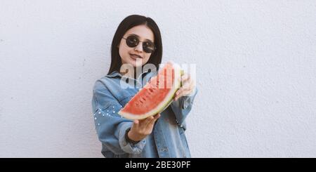 Ritratto di una bella giovane donna che mangia anguria all'aperto. Una bambina in denim trendy e occhiali da sole tenendo in mano una fetta grande di fre dolce Foto Stock