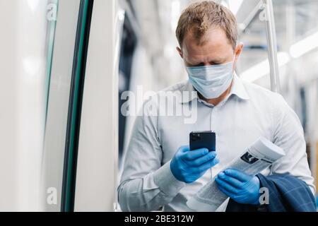 L'elegante uomo adulto in camicia bianca indossa maschera medica protettiva e guanti in gomma, controlla i newsfeed sullo smartphone, tiene il giornale, posa in tran pubblico Foto Stock