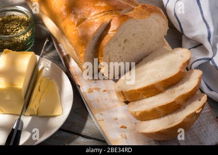 Pane affettato su tagliere con burro ed erbe. Vista dall'alto. Luce del sole e ombra frammentata. Foto Stock