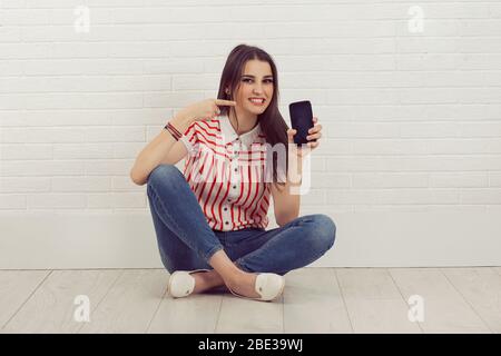 Giovane donna che punta il dito verso lo smartphone nero a schermo vuoto, seduta a gambe incrociate, guardando fotocamera, isolato bianco muro sfondo. Viso umano Foto Stock