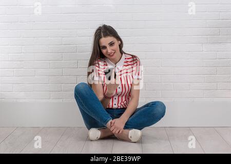 Giovane donna con cellulare, seduta a gambe incrociate sul pavimento, ottenere buone notizie sul telefono cellulare. Guardando al cellulare. Muro isolato bianco di mattoni retro gr Foto Stock