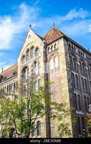 Il bellissimo edificio storico dell'Archivio Nazionale Ungherese nella capitale ungherese Budapest. L'esterno della casa circondata da alberi su una foto verticale con cielo blu sopra. Foto Stock