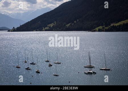 I gommoni a vela ormeggiati si trovano in mezzo ad acqua scintillante sulla superficie del Lago di Rèsia (Reschensee), in Italia, a fine estate con la montagna coperta di neve pe Foto Stock