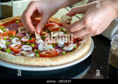 Pizza fatta in casa con verdure. Il processo di preparazione della pizza a casa. Mani di donna che fanno la pizza e che mette i funghi sulla parte superiore. Organizzazione degli ingredienti Foto Stock