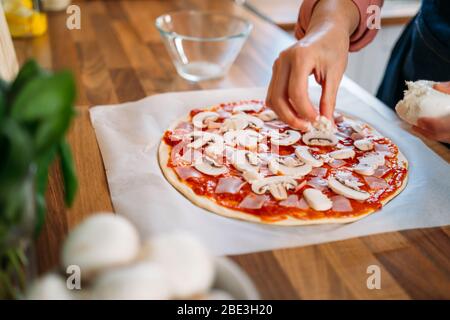 Mani femminili che aggiungono funghi ad una tradizionale pizza margarita. Preparazione di una pizza italiana originale Foto Stock