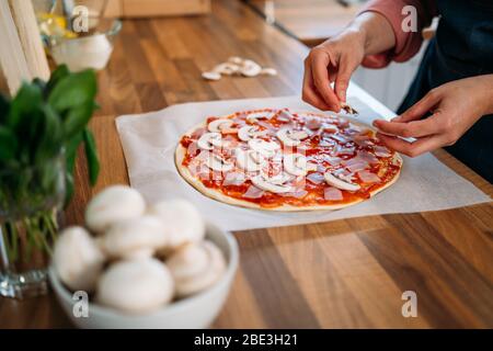 Concetto di cibo. Pizza italiana fresca cruda originale, preparazione tradizionale. Fare pizza mani femminili aggiungere funghi alla pizza, cucinare la pizza in un vero Foto Stock